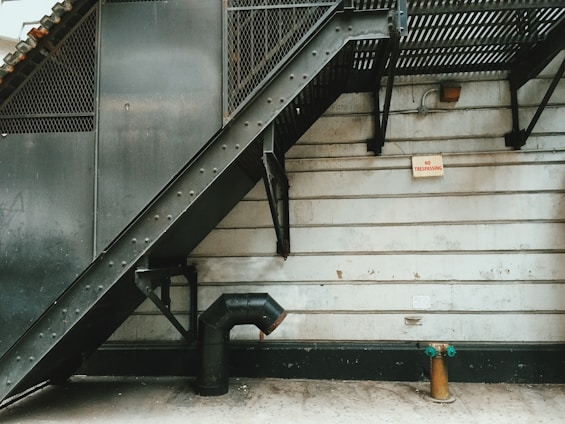 A metal fire escape staircase descends along the side of a concrete building. The structure features industrial design elements, such as rivets and metal mesh. A pipe extends from the wall near the ground, next to a fire hydrant with green caps. A 'No Trespassing' sign is mounted on the wall.