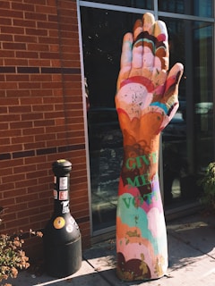 A colorful, oversized sculpture resembling a human hand stands against a red brick wall. The hand is painted in vibrant shades of pink, orange, blue, and other colors. Visible on the sculpture, the words 'GIVE ME A VOTE' are painted in green. Next to the sculpture is a black cylindrical object, possibly a cigarette disposal bin, adorned with various stickers and positioned near the glass windows of a building.