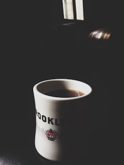 A white coffee mug with black text reading 'BROOKLYN' and a logo on the front is partially filled with coffee. The background is dimly lit, with a small amount of light coming through a window, creating a cozy and intimate atmosphere.