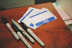 A collection of name tags and markers placed on a wooden surface. The name tags are blank, with a bold blue and white design, reading 'Hello my name is'. Four markers of varying sizes and brands are scattered nearby, with some showing a 'Super Permanent Ink' label.