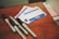 A collection of name tags and markers placed on a wooden surface. The name tags are blank, with a bold blue and white design, reading 'Hello my name is'. Four markers of varying sizes and brands are scattered nearby, with some showing a 'Super Permanent Ink' label.