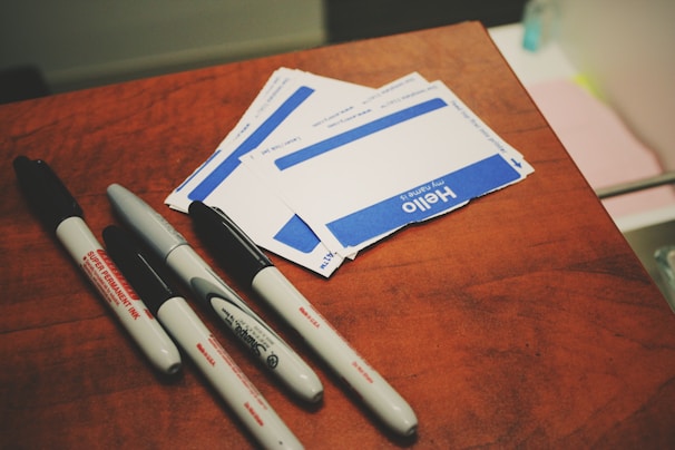 A collection of name tags and markers placed on a wooden surface. The name tags are blank, with a bold blue and white design, reading 'Hello my name is'. Four markers of varying sizes and brands are scattered nearby, with some showing a 'Super Permanent Ink' label.