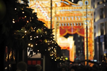 A festive and brightly lit street scene features colorful archways adorned with intricate light patterns and dark green holiday garlands with string lights and baubles in the foreground. The lighting offers a warm and inviting atmosphere, suggesting a holiday celebration or event taking place.