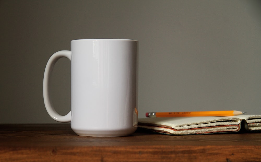 white ceramic mug beside orange pencil on open book page