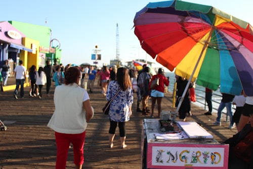 A busy boardwalk with people walking along a wooden path. There is a colorful beach umbrella providing shade to a small booth, and various brightly painted buildings are visible on the left side. The area is bustling with people enjoying the seaside atmosphere.