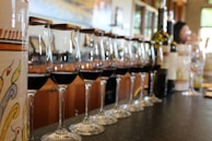 A row of wine glasses filled with red wine is neatly aligned on a countertop. Behind the glasses, a few wine bottles can be seen, and a decorative ceramic pot is partially visible on the left side of the image. In the blurred background, the setting appears to be a modern tasting room with wooden accents and a person standing in front of a window.