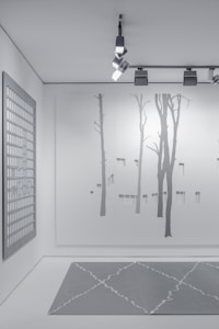 A minimalist interior space featuring art and design elements. The wall displays a large minimalist mural of tree silhouettes, painted in subtle shades of gray. An overhead track lighting system with modern fixtures illuminates the space. A grid-patterned artwork hangs on a side wall, contributing to the contemporary aesthetic. The floor features a geometric-patterned rug in muted gray tones.