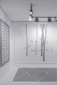 A minimalist interior space featuring art and design elements. The wall displays a large minimalist mural of tree silhouettes, painted in subtle shades of gray. An overhead track lighting system with modern fixtures illuminates the space. A grid-patterned artwork hangs on a side wall, contributing to the contemporary aesthetic. The floor features a geometric-patterned rug in muted gray tones.