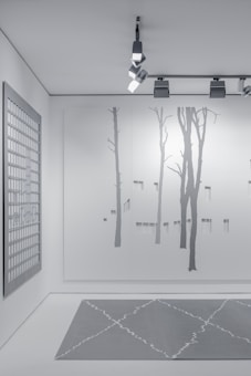 A minimalist interior space featuring art and design elements. The wall displays a large minimalist mural of tree silhouettes, painted in subtle shades of gray. An overhead track lighting system with modern fixtures illuminates the space. A grid-patterned artwork hangs on a side wall, contributing to the contemporary aesthetic. The floor features a geometric-patterned rug in muted gray tones.