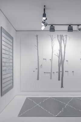 A minimalist interior space featuring art and design elements. The wall displays a large minimalist mural of tree silhouettes, painted in subtle shades of gray. An overhead track lighting system with modern fixtures illuminates the space. A grid-patterned artwork hangs on a side wall, contributing to the contemporary aesthetic. The floor features a geometric-patterned rug in muted gray tones.
