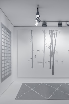 A minimalist interior space featuring art and design elements. The wall displays a large minimalist mural of tree silhouettes, painted in subtle shades of gray. An overhead track lighting system with modern fixtures illuminates the space. A grid-patterned artwork hangs on a side wall, contributing to the contemporary aesthetic. The floor features a geometric-patterned rug in muted gray tones.