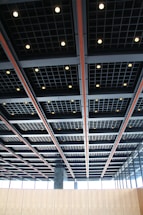 A modern architectural ceiling with a grid pattern, featuring recessed square lighting fixtures evenly spaced throughout the structure. The design is industrial and sleek, incorporating contrasting colors with black, gray, and orange elements. Large windows are visible along the walls, allowing natural light to complement the interior lighting.