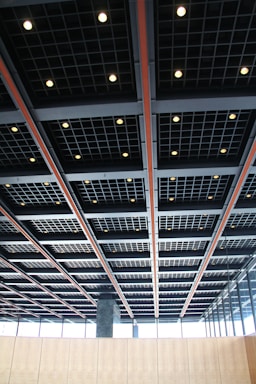 A modern architectural ceiling with a grid pattern, featuring recessed square lighting fixtures evenly spaced throughout the structure. The design is industrial and sleek, incorporating contrasting colors with black, gray, and orange elements. Large windows are visible along the walls, allowing natural light to complement the interior lighting.