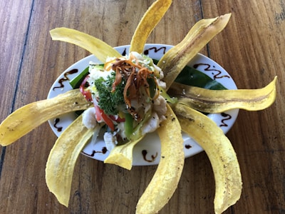 A beautifully arranged dish with long strips of fried plantains fanning out around a central pile of fresh vegetables, cheese, and herbs. The plate includes colorful ingredients like chopped red and green peppers, cilantro, and a hint of grated cheese, all garnished with a drizzle of dark sauce around the edge. The setting rests on a rustic wooden table.