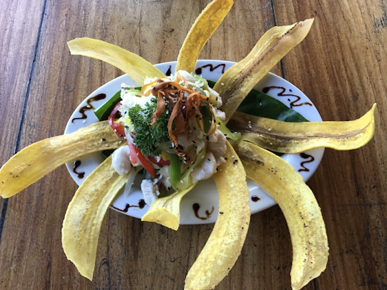 A beautifully arranged dish with long strips of fried plantains fanning out around a central pile of fresh vegetables, cheese, and herbs. The plate includes colorful ingredients like chopped red and green peppers, cilantro, and a hint of grated cheese, all garnished with a drizzle of dark sauce around the edge. The setting rests on a rustic wooden table.