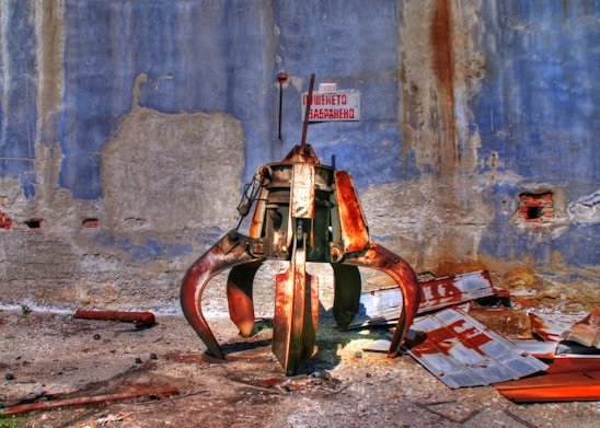 A large, rusted industrial claw machine is positioned against a weathered blue and gray wall, surrounded by scattered metal debris. The environment appears to be abandoned or neglected, with peeling paint and crumbling textures on the wall.