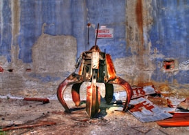 A large, rusted industrial claw machine is positioned against a weathered blue and gray wall, surrounded by scattered metal debris. The environment appears to be abandoned or neglected, with peeling paint and crumbling textures on the wall.
