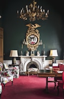 An elegantly decorated room featuring a vintage chandelier hanging from the ceiling, a large ornate mirror with a golden eagle motif above a white marble fireplace. The room has lush, patterned armchairs and a vibrant red carpet. Flanking the fireplace are two lamps with large shades, and the dark green walls add sophistication. The room also includes a wooden table with decorative items.