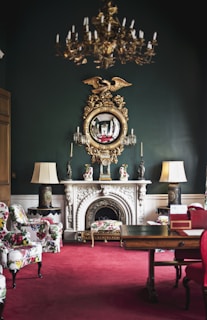An elegantly decorated room featuring a vintage chandelier hanging from the ceiling, a large ornate mirror with a golden eagle motif above a white marble fireplace. The room has lush, patterned armchairs and a vibrant red carpet. Flanking the fireplace are two lamps with large shades, and the dark green walls add sophistication. The room also includes a wooden table with decorative items.