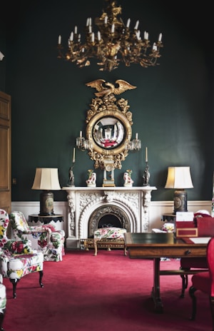 An elegantly decorated room featuring a vintage chandelier hanging from the ceiling, a large ornate mirror with a golden eagle motif above a white marble fireplace. The room has lush, patterned armchairs and a vibrant red carpet. Flanking the fireplace are two lamps with large shades, and the dark green walls add sophistication. The room also includes a wooden table with decorative items.