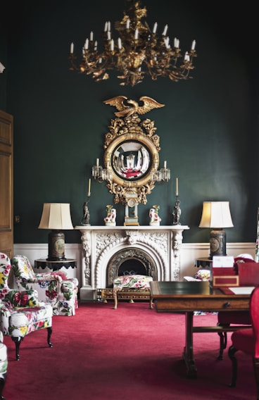 An elegantly decorated room featuring a vintage chandelier hanging from the ceiling, a large ornate mirror with a golden eagle motif above a white marble fireplace. The room has lush, patterned armchairs and a vibrant red carpet. Flanking the fireplace are two lamps with large shades, and the dark green walls add sophistication. The room also includes a wooden table with decorative items.