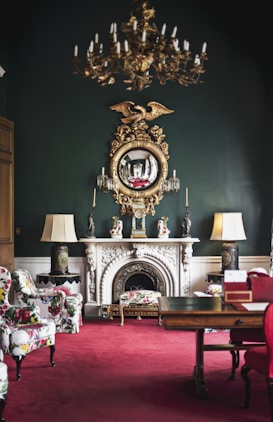 An elegantly decorated room featuring a vintage chandelier hanging from the ceiling, a large ornate mirror with a golden eagle motif above a white marble fireplace. The room has lush, patterned armchairs and a vibrant red carpet. Flanking the fireplace are two lamps with large shades, and the dark green walls add sophistication. The room also includes a wooden table with decorative items.
