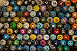 A collection of spray paint cans arranged closely together, showcasing a variety of vibrant colors such as blues, greens, reds, oranges, and yellows. The arrangement is from a top-down perspective, displaying the caps and nozzles of each can.
