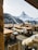 A wooden outdoor restaurant terrace with rustic tables and benches is covered with soft fur throws. It overlooks a breathtaking view of snow-capped mountains and evergreen forests, with a prominent peak in the background under a bright, clear sky.