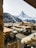 A wooden outdoor restaurant terrace with rustic tables and benches is covered with soft fur throws. It overlooks a breathtaking view of snow-capped mountains and evergreen forests, with a prominent peak in the background under a bright, clear sky.