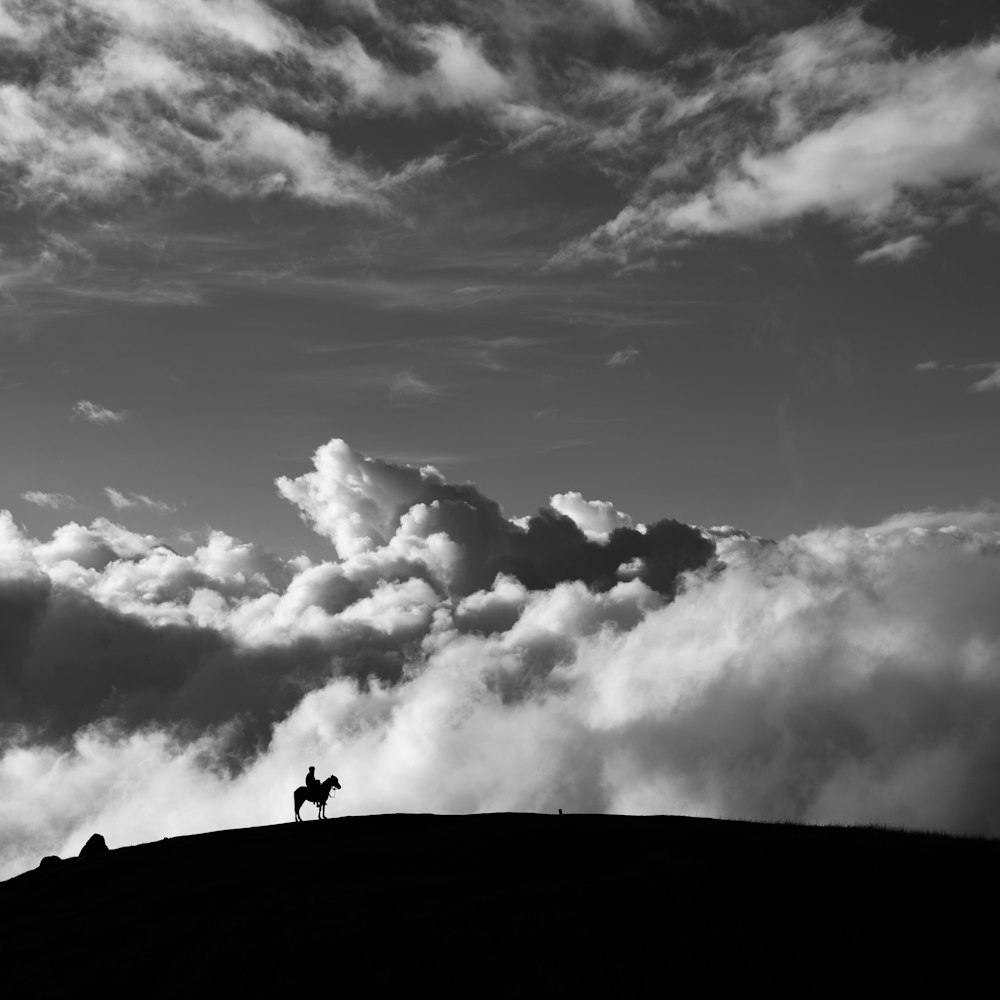 적운 구름을 배경으로 산에서 말을 타고 있는 남자의 실루엣의 회색조 사진