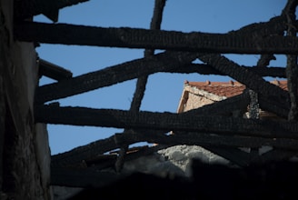 A view through the charred remains of a roof structure, with burnt wooden beams creating a dark, jagged frame against a clear blue sky. In the background, the roof of a partially damaged building with red tiles is visible.