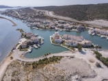 Aerial view of a coastal residential area with houses surrounded by water channels and moored boats. The landscape includes hilly terrain at the background and a winding road cutting through the area. Vegetation and open land are visible around the buildings.