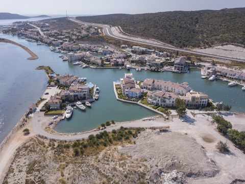 Aerial view of a coastal residential area with houses surrounded by water channels and moored boats. The landscape includes hilly terrain at the background and a winding road cutting through the area. Vegetation and open land are visible around the buildings.