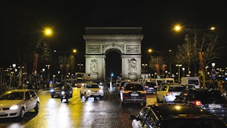 A nighttime scene of the busy Champs-Élysées in Paris, filled with cars under bright streetlights. The iconic Arc de Triomphe stands prominently in the background, framed by leafless trees. Headlights and taillights create reflections on the wet cobblestone street, contributing to the vibrant city atmosphere.