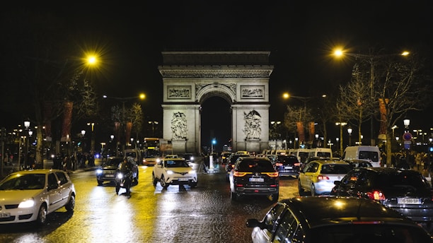 A nighttime scene of the busy Champs-Élysées in Paris, filled with cars under bright streetlights. The iconic Arc de Triomphe stands prominently in the background, framed by leafless trees. Headlights and taillights create reflections on the wet cobblestone street, contributing to the vibrant city atmosphere.