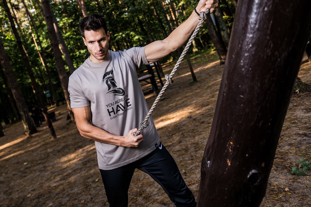 man pulling gray rope beside tree trunk in forest