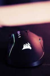 A sleek, black gaming mouse with a smooth, ergonomic design. The mouse features backlit accents, including a glowing logo on its surface. The lighting gives off a tech-savvy, modern feel.