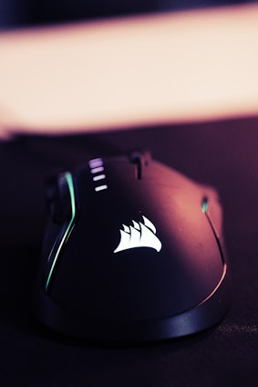 A sleek, black gaming mouse with a smooth, ergonomic design. The mouse features backlit accents, including a glowing logo on its surface. The lighting gives off a tech-savvy, modern feel.
