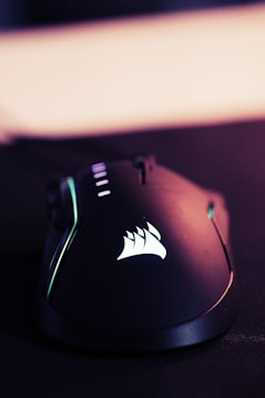 A sleek, black gaming mouse with a smooth, ergonomic design. The mouse features backlit accents, including a glowing logo on its surface. The lighting gives off a tech-savvy, modern feel.