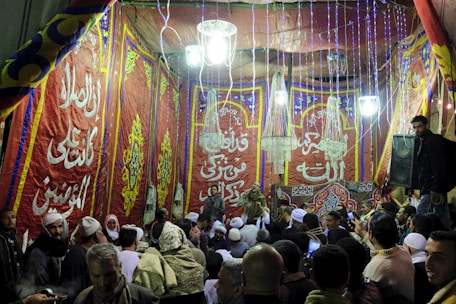 A gathering of people is seen inside a decorated tent with vibrant red, blue, and yellow patterns and Arabic calligraphy on the walls. Several individuals are wearing traditional clothing with some filming or taking pictures using their phones. The scene is illuminated by chandeliers and string lights hanging from above.