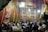 A gathering of people is seen inside a decorated tent with vibrant red, blue, and yellow patterns and Arabic calligraphy on the walls. Several individuals are wearing traditional clothing with some filming or taking pictures using their phones. The scene is illuminated by chandeliers and string lights hanging from above.