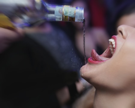 A person tilts their head back with an open mouth as liquid is poured from a bottle above. The scene suggests a lively, possibly party-related atmosphere.