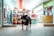 A dog wearing a black jacket stands at the entrance of a brightly lit and colorful store. The store interior contains various products on shelves, and a person is visible in the background.