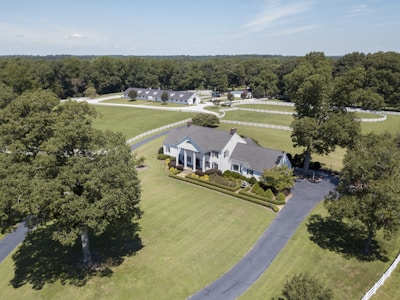 Aerial view of a large, elegant country estate surrounded by lush green lawns and tall trees. The property features a sizable, classic white house with a gray roof, accompanied by immaculately trimmed hedges and additional buildings visible in the background. A long, winding driveway leads to the front of the house, and a white fence borders the expansive grounds.