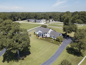 Aerial view of a large, elegant country estate surrounded by lush green lawns and tall trees. The property features a sizable, classic white house with a gray roof, accompanied by immaculately trimmed hedges and additional buildings visible in the background. A long, winding driveway leads to the front of the house, and a white fence borders the expansive grounds.