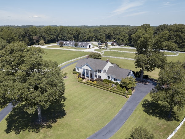 Aerial view of a large, elegant country estate surrounded by lush green lawns and tall trees. The property features a sizable, classic white house with a gray roof, accompanied by immaculately trimmed hedges and additional buildings visible in the background. A long, winding driveway leads to the front of the house, and a white fence borders the expansive grounds.