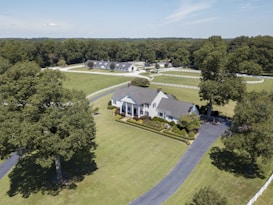 Aerial view of a large, elegant country estate surrounded by lush green lawns and tall trees. The property features a sizable, classic white house with a gray roof, accompanied by immaculately trimmed hedges and additional buildings visible in the background. A long, winding driveway leads to the front of the house, and a white fence borders the expansive grounds.
