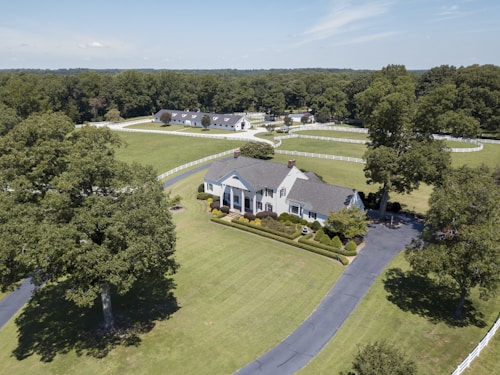 Aerial view of a large, elegant country estate surrounded by lush green lawns and tall trees. The property features a sizable, classic white house with a gray roof, accompanied by immaculately trimmed hedges and additional buildings visible in the background. A long, winding driveway leads to the front of the house, and a white fence borders the expansive grounds.