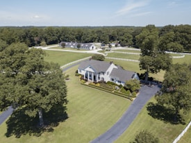 Aerial view of a large, elegant country estate surrounded by lush green lawns and tall trees. The property features a sizable, classic white house with a gray roof, accompanied by immaculately trimmed hedges and additional buildings visible in the background. A long, winding driveway leads to the front of the house, and a white fence borders the expansive grounds.