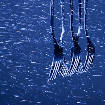 Four shiny metal forks are positioned vertically with the tines facing downward, illuminated and contrasted against a backdrop of dynamic, blurred streaks of light that resemble rainfall or streaking motion.